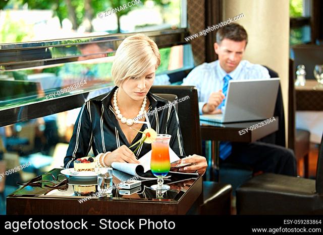 Young woman sitting at table in cafe, reading magazine. Businessman using laptop in the background