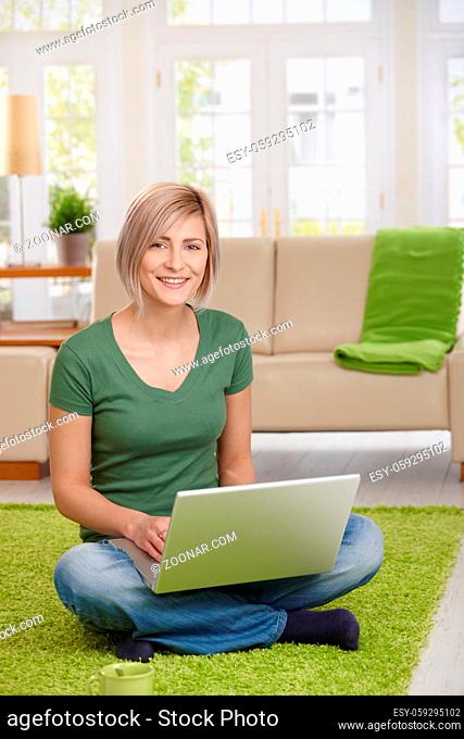 Happy woman sitting on floor at home in living room using laptop computer, looking at camera