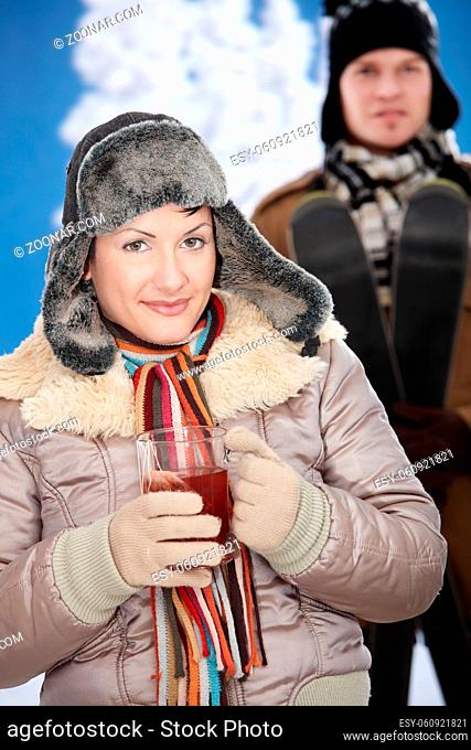 Young couple in snow at winter wearing warm clothes, smiling. Woman drinking cup of hot tea
