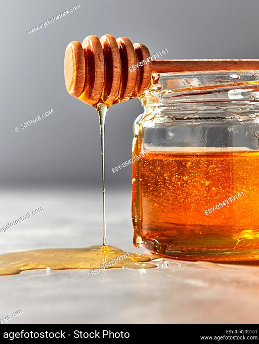 Dripping sweet flower honey from dipper above the glass pot with dessert on a gray stone background, copy space. Traditional healthy sweetness