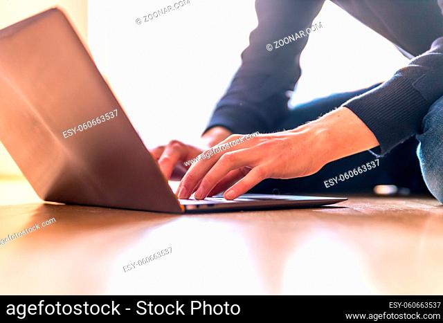 Young man with a laptop is working on the wooden floor, light comes in from the window