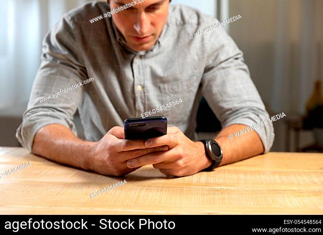 Front view close up of a young Caucasian man sitting at a table using a smartphone at home in the evening
