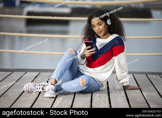 Pretty ethnic girl in stylish casual outfit using headphones with phone and looking flirty at camera