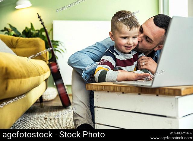 Father kissing son using laptop in living room at home