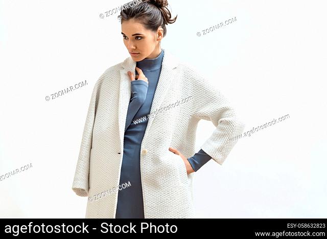 Sexy girl is posing in the studio on the light background. She wears a gray dress and a white coat. Woman holds left hand in the pocket