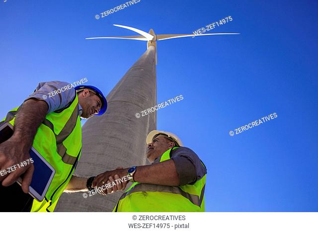 Low angle view of technicians shaking hands in front of wind turbine
