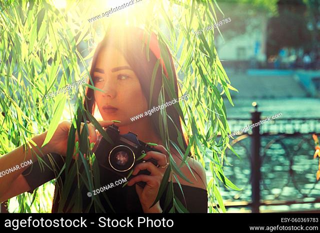 Asian Girl with camera in the park in sunset time. Brunette girl with film camera in the vintage colored image posing against willow branches and looking away