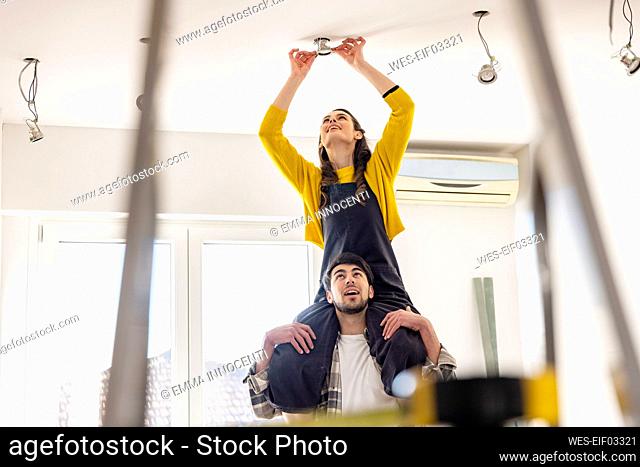 Young woman sitting on boyfriend shoulders installing light bulb in new home