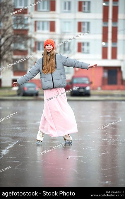 Girl teenager skates on ice in the city.Young girl in a long dress and a warm jacket on the ice