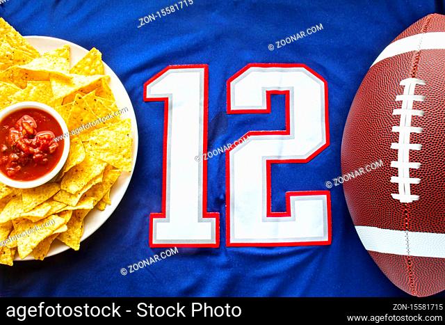 An American football with organic nacho chips and mild salsa on a white blue football jersey with the 12 number on horizontal view
