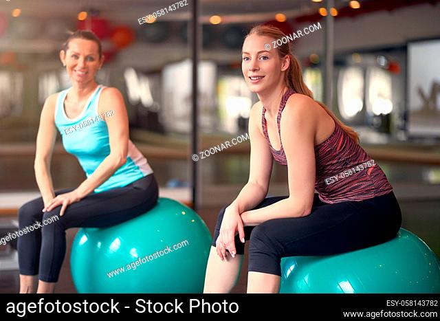 Two healthy young women with pilates or gym balls taking a break from their workout in a gym sitting smiling at the camera in an active lifestyle and fitness...