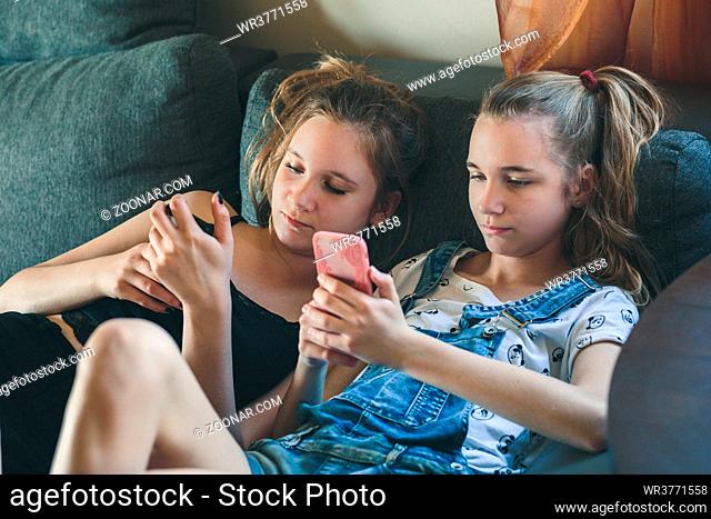 Young women using mobile phones watching music clip, texting, messaging. Teenagers using the smartphones, sitting on sofa at home