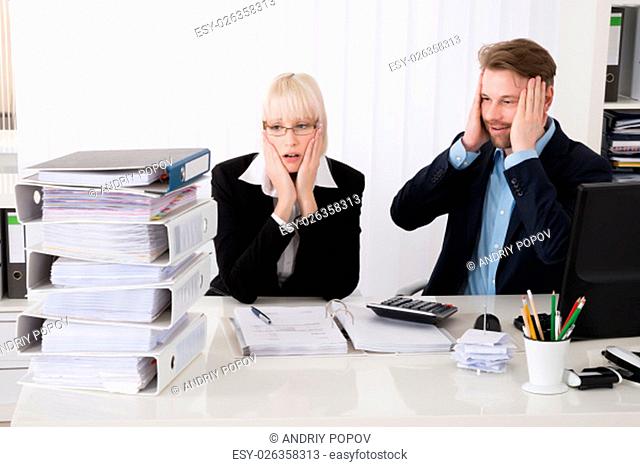 Stressed Young Businesspeople Looking At Stack Of Folders In Office