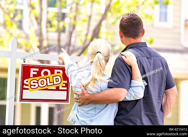 Caucasian couple facing and pointing to front of sold real estate sign and house