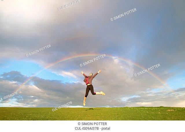 Young female hiker jumping mid air below rainbow