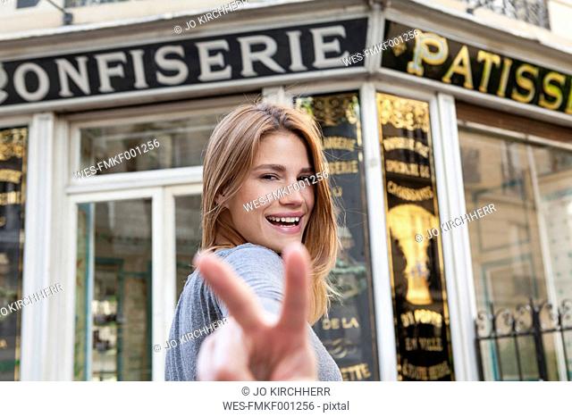 France, Paris, portrait of young woman showing victory sign in front of pastry shop
