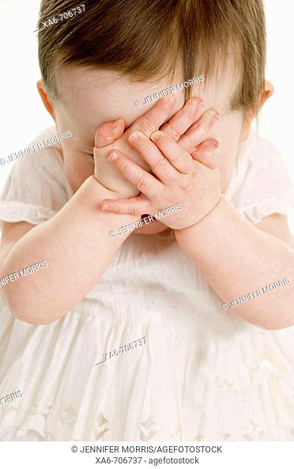 A one-year-old girl wearing a white dress clasps her hands over here face