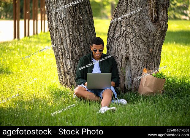 Outdoor pastime. Young dark-skinned man smiling into laptop sitting under tree on grass near bag with snack on summer day