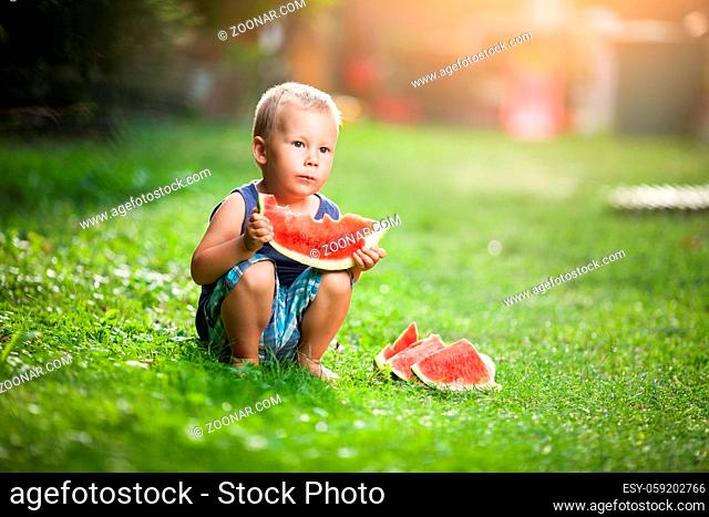Cute toddler sitting outdoords and eating a slice of watermelon