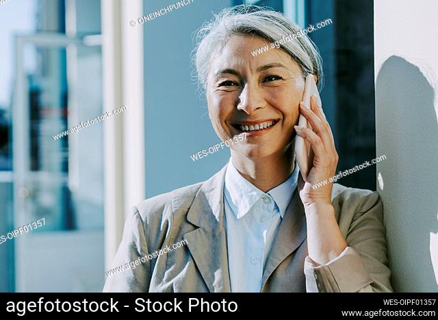 Happy mature woman talking on mobile phone by wall