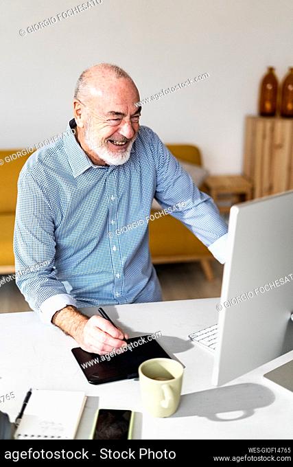 Cheerful senior businessman with graphic tablet looking at computer in home office