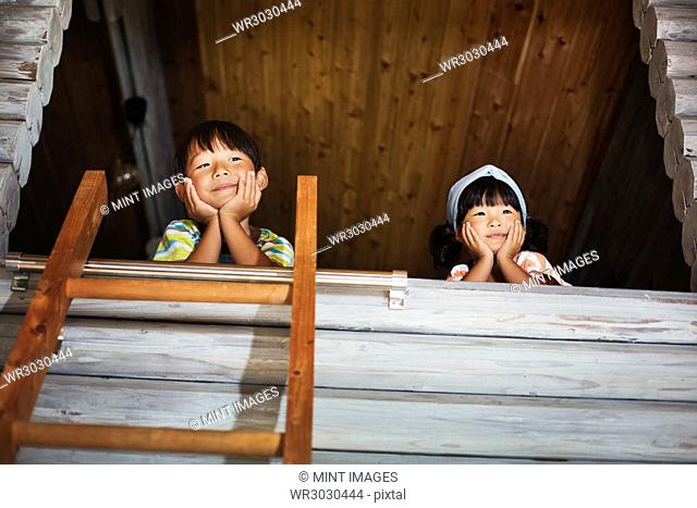 Low angle view of boy and young girl with black hair looking out of window, ladder leaning against wooden wall