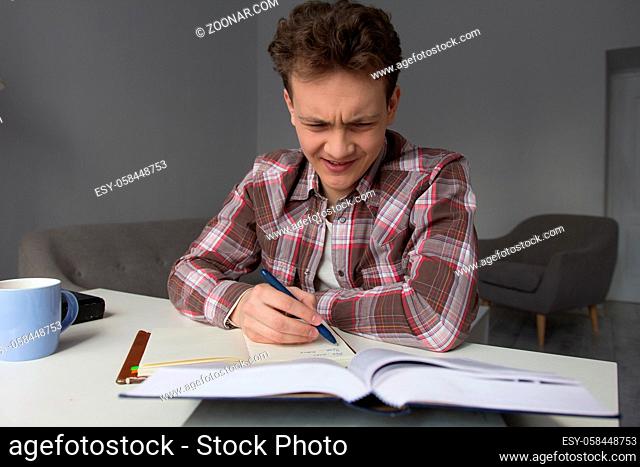 Guy attempting to do his homework. Young good looking boy with lcurly hair studying at home in living room with notebook and book with confused face expression