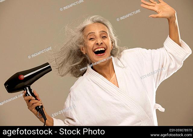 A happy old woman drying her hair with a blow-dryer
