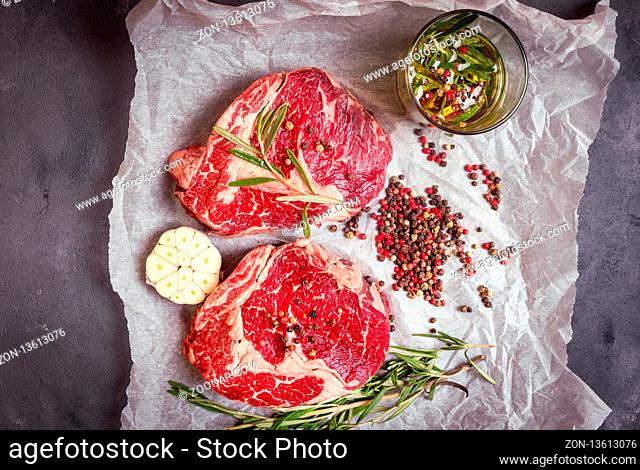 Raw juicy steaks with seasonings on a baking paper ready for roasting on rustic concrete background. Fresh marbled meat steaks with herbs, garlic, olive oil
