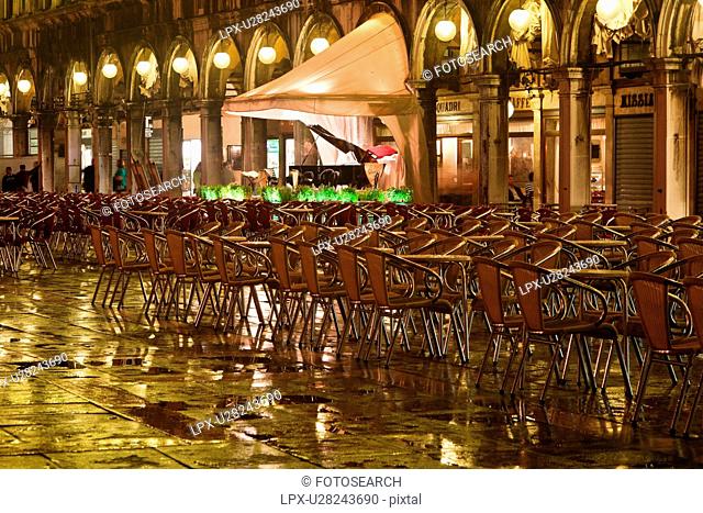 Piazza San Marco at night, with Aqua Alta flooding, lights of loggia reflected in puddles, rows of empty red and yellow chairs, empty orchestra