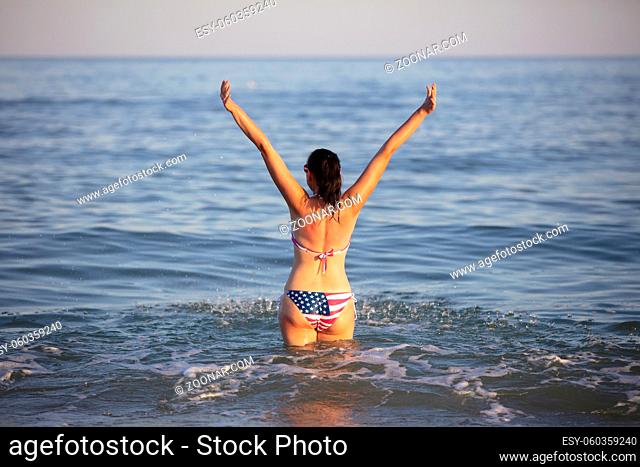 A middle-aged woman stands in sea water with her arms raised in a swimsuit with an American flag print
