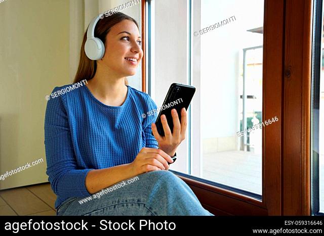 Young woman listening to music with headphones on floor looking through the window