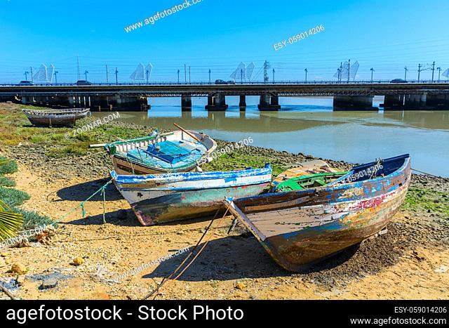 Old fishing boats on the shore of a river, with a bridge in the background