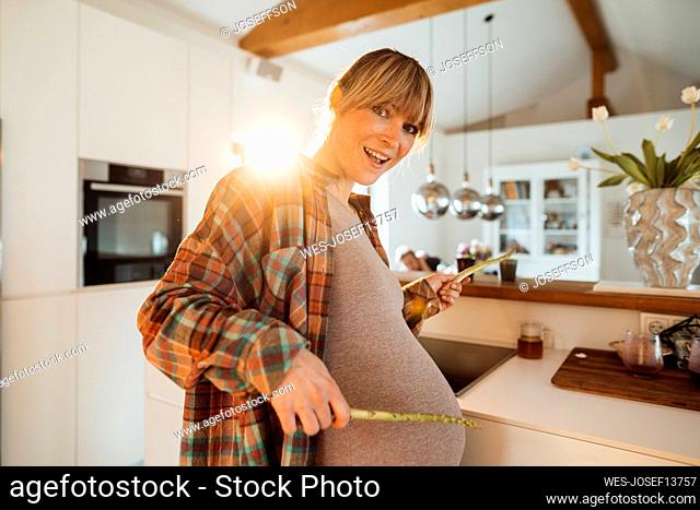 Happy expectant woman playing with asparagus sticks in kitchen at home