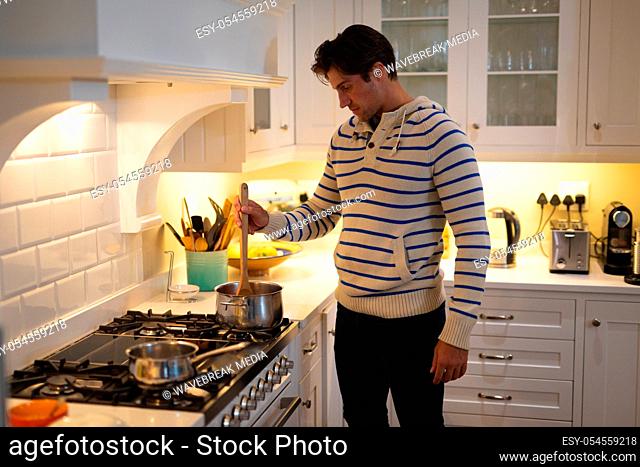 Side view of a Caucasian man at home wearing a blue striped sweater, standing in the kitchen and cooking, stirring a pan on the hob