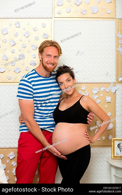 Beautiful young couple expecting baby soon. Husband touching his wife's belly and they both looking at camera at home