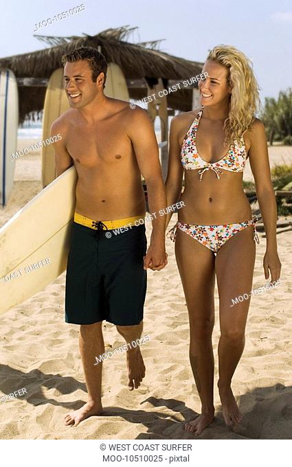 Surfer couple holding hands carrying surfboard walking on beach