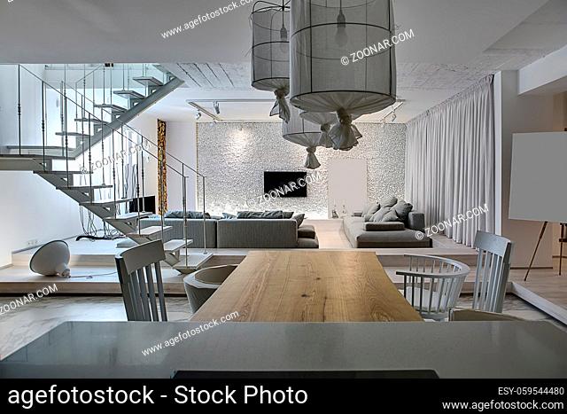 Modern interior with white walls. There is a white wooden stair with a metal railing, gray sofas with pillows, glowing lamps, tables with chairs, TV