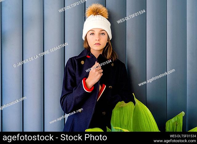 Serious teenage girl wearing winter coat standing by houseplant against wall at home