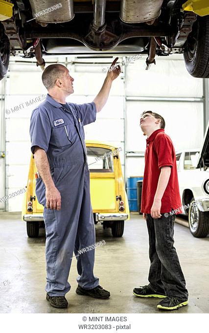 A Caucasian senior male car mechanic talks to his grandson about car repairs in a classic car repair shop