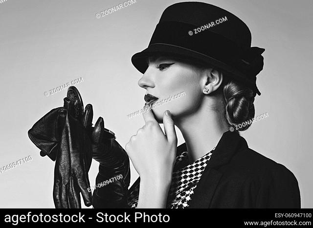 Beautiful young woman with red lips make-up in retro black and white dress, hat, jacket and leather gloves standing on grey background looking at pocket mirror