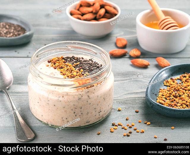 Overnight oats in jar and ingredients - chia seeds, almond, honey and pollen on gray wooden table background. Healthy breakfast oatmeal recipe idea