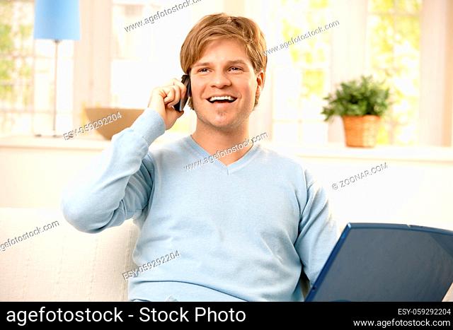 Smiling young man using cellphone, holding computer, sitting on couch in living room