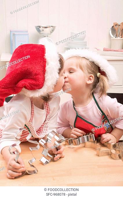 Two small girls in Father Christmas hats kissing in kitchen