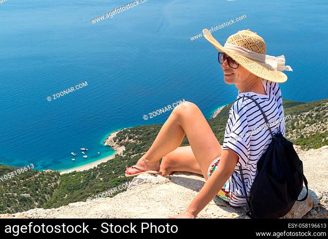 Active sporty woman on summer vacations sitting on old stone wall at Lubenice village, wearing straw hat and beach backpack enjoying beautiful coastal view of...
