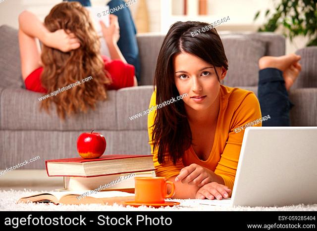 Teen girls studying at home in living room with books and laptop, girl in front looking at camera