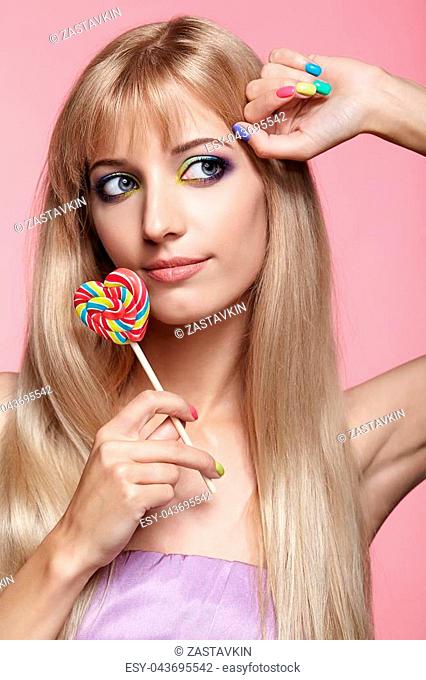 Beauty portrait of young blonde woman on pink background. Female with candy lollipop on stick in hands. Girl has finger nails with bright yellow, green
