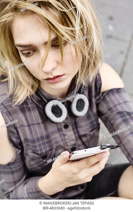 Fashionably dressed young woman, teenager, sitting depressed on the floor with smartphone in her hand, North Rhine-Westphalia, Germany, Europe