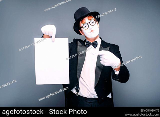 Male mime actor with empty paper sheet. Pantomime in suit, gloves, glasses, make-up mask and hat. April fools day concept