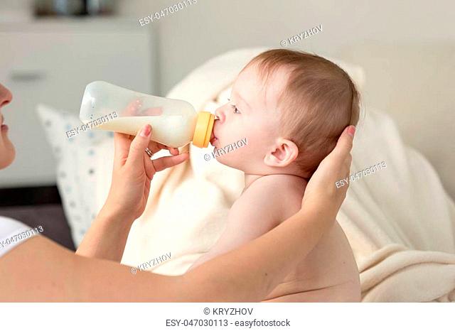 Closeup photo of mother giving milk from bottle to her baby son sitting on sofa